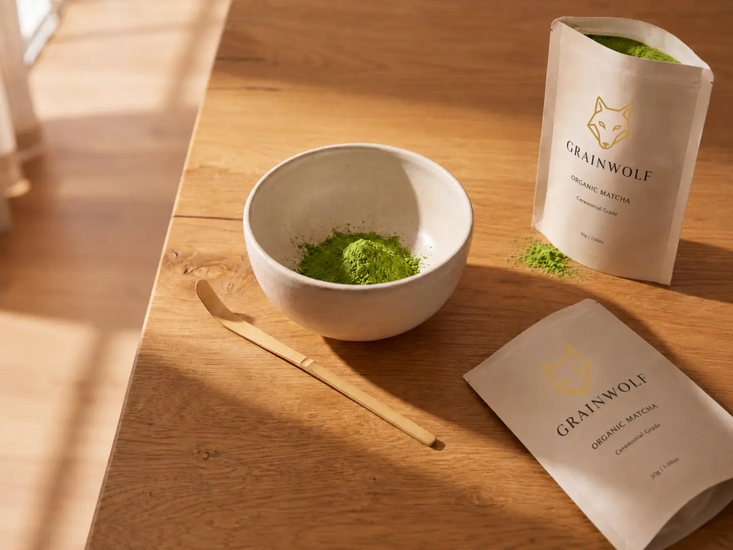Grain Wolf organic ceremonial matcha green tea powder displayed in a white ceramic bowl next to a traditional bamboo scoop and two branded kraft pouches on a sunlit wooden table.